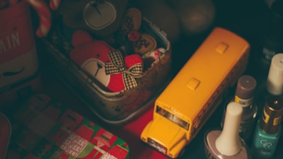 The image contains a collection of items including a yellow toy school bus, a tin containing stuffed toy bears, bottles of nail polish, and a festive gift box. The scene is dimly lit, creating a cozy, nostalgic atmosphere.