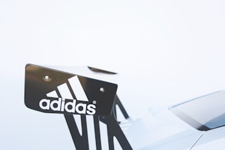 A close-up of a car's rear spoiler featuring the Adidas logo. The logo is white and contrasts with the dark color of the spoiler. The background is a soft, light color, creating a minimalist and clean appearance.