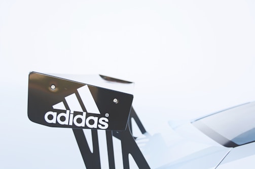 A close-up of a car's rear spoiler featuring the Adidas logo. The logo is white and contrasts with the dark color of the spoiler. The background is a soft, light color, creating a minimalist and clean appearance.