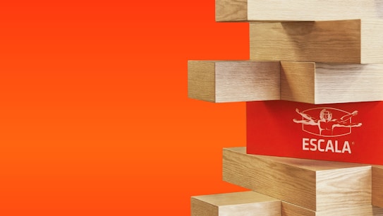 A series of wooden blocks are stacked in a staggered pattern against a vibrant orange background. One of the blocks has a red face with the word 'ESCALA' and a stylized human figure printed on it.