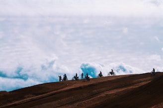 A group of motorcyclists ride along a dirt ridge with a backdrop of expansive, cloudy skies. The silhouettes of the riders are visible against the bright sky, emphasizing the adventurous and daring atmosphere.