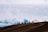 A group of motorcyclists ride along a dirt ridge with a backdrop of expansive, cloudy skies. The silhouettes of the riders are visible against the bright sky, emphasizing the adventurous and daring atmosphere.