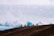 A group of motorcyclists ride along a dirt ridge with a backdrop of expansive, cloudy skies. The silhouettes of the riders are visible against the bright sky, emphasizing the adventurous and daring atmosphere.