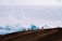 A group of motorcyclists ride along a dirt ridge with a backdrop of expansive, cloudy skies. The silhouettes of the riders are visible against the bright sky, emphasizing the adventurous and daring atmosphere.