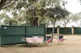 A picnic area is set in a park with a wooden green fence marked 'A5'. Two folding chairs adorned with American flag patterns are positioned next to a picnic table. The table is covered with a red and white checkered tablecloth, hosting picnic items, including a closed cooler. The setting is surrounded by trees, suggesting a quiet, outdoor environment.