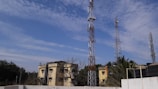 Multiple telecommunications towers are set against a partly cloudy blue sky. Two older yellow buildings are situated in front, surrounded by some trees and vegetation. The scene suggests an urban or semi-urban environment.