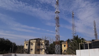 Multiple telecommunications towers are set against a partly cloudy blue sky. Two older yellow buildings are situated in front, surrounded by some trees and vegetation. The scene suggests an urban or semi-urban environment.