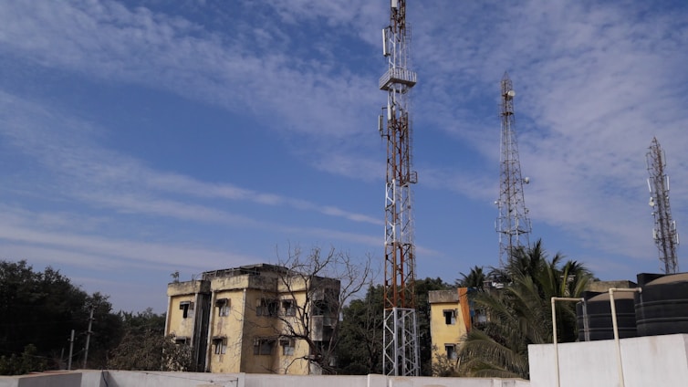 Multiple telecommunications towers are set against a partly cloudy blue sky. Two older yellow buildings are situated in front, surrounded by some trees and vegetation. The scene suggests an urban or semi-urban environment.