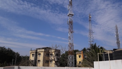 Multiple telecommunications towers are set against a partly cloudy blue sky. Two older yellow buildings are situated in front, surrounded by some trees and vegetation. The scene suggests an urban or semi-urban environment.
