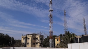 Multiple telecommunications towers are set against a partly cloudy blue sky. Two older yellow buildings are situated in front, surrounded by some trees and vegetation. The scene suggests an urban or semi-urban environment.