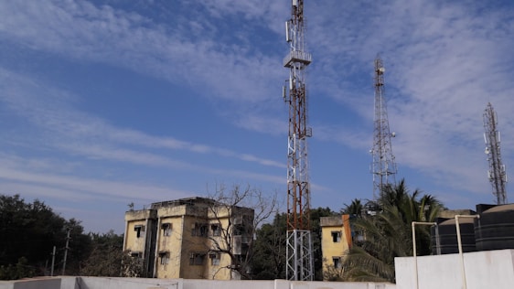 Multiple telecommunications towers are set against a partly cloudy blue sky. Two older yellow buildings are situated in front, surrounded by some trees and vegetation. The scene suggests an urban or semi-urban environment.