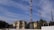 Multiple telecommunications towers are set against a partly cloudy blue sky. Two older yellow buildings are situated in front, surrounded by some trees and vegetation. The scene suggests an urban or semi-urban environment.