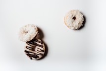 Three donuts are arranged on a white surface. Two of them are covered in white shredded coconut, while the third is decorated with chocolate drizzle.