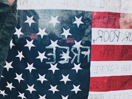 An American flag is displayed, featuring stars and stripes. Text is faintly visible on the white stripes, with the word 'FREE' prominently written in reverse on the blue field with stars. The edge of a person's arm draped in a dark sleeve is visible on the left.
