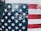 An American flag is displayed, featuring stars and stripes. Text is faintly visible on the white stripes, with the word 'FREE' prominently written in reverse on the blue field with stars. The edge of a person's arm draped in a dark sleeve is visible on the left.