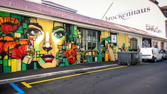 A vibrant mural adorns the side of a building, featuring a stylized portrait of a woman's face surrounded by geometric shapes and vivid colors. Bright red flowers and abstract patterns are interspersed in the artwork. The building, labeled 'Zurcher Brockenhaus,' is situated along a paved street with a couple of parked vehicles and a row of garbage bins nearby.