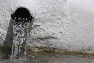 Water flows out of a round pipe embedded in a textured, white wall. The water appears clear as it pours out, creating ripples and splashes as it hits the surface below.