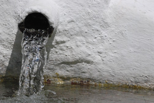 Water flows out of a round pipe embedded in a textured, white wall. The water appears clear as it pours out, creating ripples and splashes as it hits the surface below.