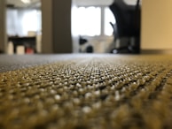 A close-up view of a textured carpet with a blurred background, suggesting an office or indoor environment. The carpet shows intricate weave patterns, and the space appears to be lit by natural light coming through a window.