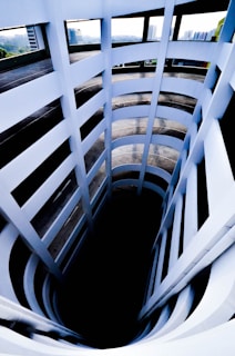 A multi-level spiral parking garage with a deep central void, surrounded by vertical white walls and horizontal ledges. The structure is partially open to the outside, with windows showing distant city buildings and greenery.