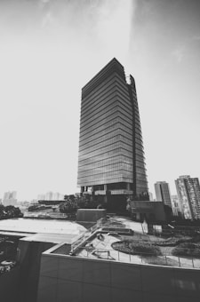 A modern skyscraper dominates the urban landscape with its sleek, rectangular design and reflective glass facade. Surrounding the building are other high-rise structures, trees, and an overlook with railings that offer a view of the city. The image is in black and white, giving it a classic and timeless feel.
