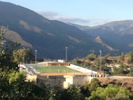 A small, elevated football field surrounded by a wire fence is nestled within a mountainous landscape. The field is brightly lit by tall floodlights and a few players can be seen on the green artificial turf. The surrounding area is densely vegetated with various types of trees and shrubs, and the backdrop of majestic mountains under a clear blue sky adds to the scenic beauty.