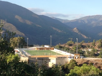 A small, elevated football field surrounded by a wire fence is nestled within a mountainous landscape. The field is brightly lit by tall floodlights and a few players can be seen on the green artificial turf. The surrounding area is densely vegetated with various types of trees and shrubs, and the backdrop of majestic mountains under a clear blue sky adds to the scenic beauty.