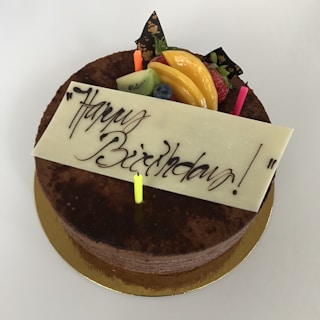 A chocolate cake with a white chocolate plaque that has 'Happy Birthday' written in elegant script. The cake is decorated with colorful candles and fresh fruits, including kiwi, peaches, strawberries, and blueberries, as well as chocolate decorations with gold accents.