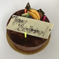 A chocolate cake with a white chocolate plaque that has 'Happy Birthday' written in elegant script. The cake is decorated with colorful candles and fresh fruits, including kiwi, peaches, strawberries, and blueberries, as well as chocolate decorations with gold accents.