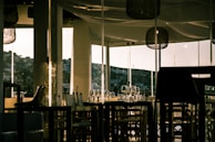 A dimly lit restaurant interior with glass walls allowing a view of a hillside in the distance. Empty chairs and tables are neatly arranged, adorned with wine glasses and tableware. The warm lighting creates an inviting ambiance, with reflections on the glass surfaces.