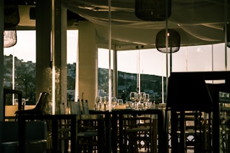 A dimly lit restaurant interior with glass walls allowing a view of a hillside in the distance. Empty chairs and tables are neatly arranged, adorned with wine glasses and tableware. The warm lighting creates an inviting ambiance, with reflections on the glass surfaces.