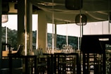 A dimly lit restaurant interior with glass walls allowing a view of a hillside in the distance. Empty chairs and tables are neatly arranged, adorned with wine glasses and tableware. The warm lighting creates an inviting ambiance, with reflections on the glass surfaces.