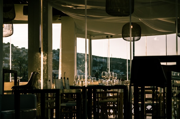 A dimly lit restaurant interior with glass walls allowing a view of a hillside in the distance. Empty chairs and tables are neatly arranged, adorned with wine glasses and tableware. The warm lighting creates an inviting ambiance, with reflections on the glass surfaces.