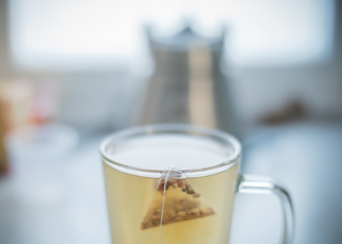 A glass cup filled with a light-colored tea, featuring a triangular tea bag partially submerged inside. The background is softly blurred, with hints of a kettle and other kitchen items.
