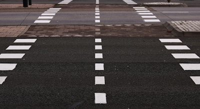 A series of white dashed lines on a dark asphalt road marking pedestrian crosswalks. The crosswalks are aligned in parallel rows with additional solid white lines on the sides, leading to sidewalks composed of interlocking pavers.