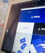 A computer screen displays a graphic design software with a brand identity project open, featuring multiple blue-themed design variations for a brand labeled 'APATIE'. The image includes different versions of the brand's logo and designs on a grid layout. The background shows a blurred bottle and some greenery out of focus.