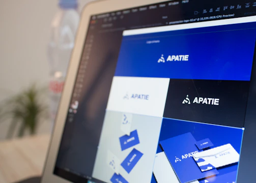 A computer screen displays a graphic design software with a brand identity project open, featuring multiple blue-themed design variations for a brand labeled 'APATIE'. The image includes different versions of the brand's logo and designs on a grid layout. The background shows a blurred bottle and some greenery out of focus.
