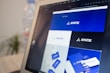 A computer screen displays a graphic design software with a brand identity project open, featuring multiple blue-themed design variations for a brand labeled 'APATIE'. The image includes different versions of the brand's logo and designs on a grid layout. The background shows a blurred bottle and some greenery out of focus.