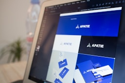 A computer screen displays a graphic design software with a brand identity project open, featuring multiple blue-themed design variations for a brand labeled 'APATIE'. The image includes different versions of the brand's logo and designs on a grid layout. The background shows a blurred bottle and some greenery out of focus.