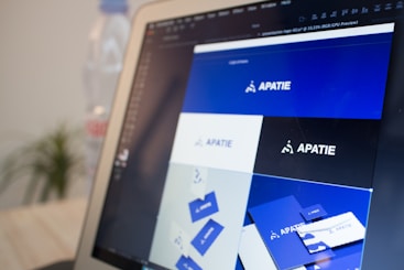 A computer screen displays a graphic design software with a brand identity project open, featuring multiple blue-themed design variations for a brand labeled 'APATIE'. The image includes different versions of the brand's logo and designs on a grid layout. The background shows a blurred bottle and some greenery out of focus.