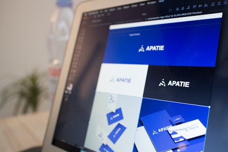 A computer screen displays a graphic design software with a brand identity project open, featuring multiple blue-themed design variations for a brand labeled 'APATIE'. The image includes different versions of the brand's logo and designs on a grid layout. The background shows a blurred bottle and some greenery out of focus.