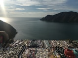 Various beaded necklaces and bracelets are laid out on a table with a scenic view of a calm sea and distant mountains in the background. The sky is partly cloudy with the sunlight creating a shimmering effect on the water.