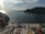 Various beaded necklaces and bracelets are laid out on a table with a scenic view of a calm sea and distant mountains in the background. The sky is partly cloudy with the sunlight creating a shimmering effect on the water.