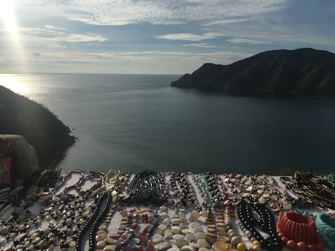 Various beaded necklaces and bracelets are laid out on a table with a scenic view of a calm sea and distant mountains in the background. The sky is partly cloudy with the sunlight creating a shimmering effect on the water.