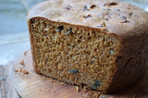 A loaf of brown bread with a piece cut out, showing its textured interior filled with seeds and grains. The crust appears golden brown and slightly rough. It rests on a wooden cutting board, with some crumbs scattered around.