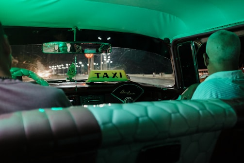 Inside the vintage car, two people are seated, one person visible from the back and another in the rearview mirror. The dashboard features a prominent 'TAXI' sign and the interior is lit with a green glow. The road ahead is dimly lit, with streetlights illuminating the night scene.