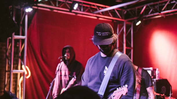 Musicians performing on stage, featuring one wearing a cap and playing an electric guitar, while another stands in the background with a microphone. The setting includes red curtains, stage lighting, and metal truss structures.