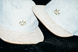 Two light-colored baseball caps with embroidered logos. The caps appear worn, with some scuffs and markings on the brims, set against a dark background.