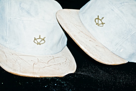 Two light-colored baseball caps with embroidered logos. The caps appear worn, with some scuffs and markings on the brims, set against a dark background.