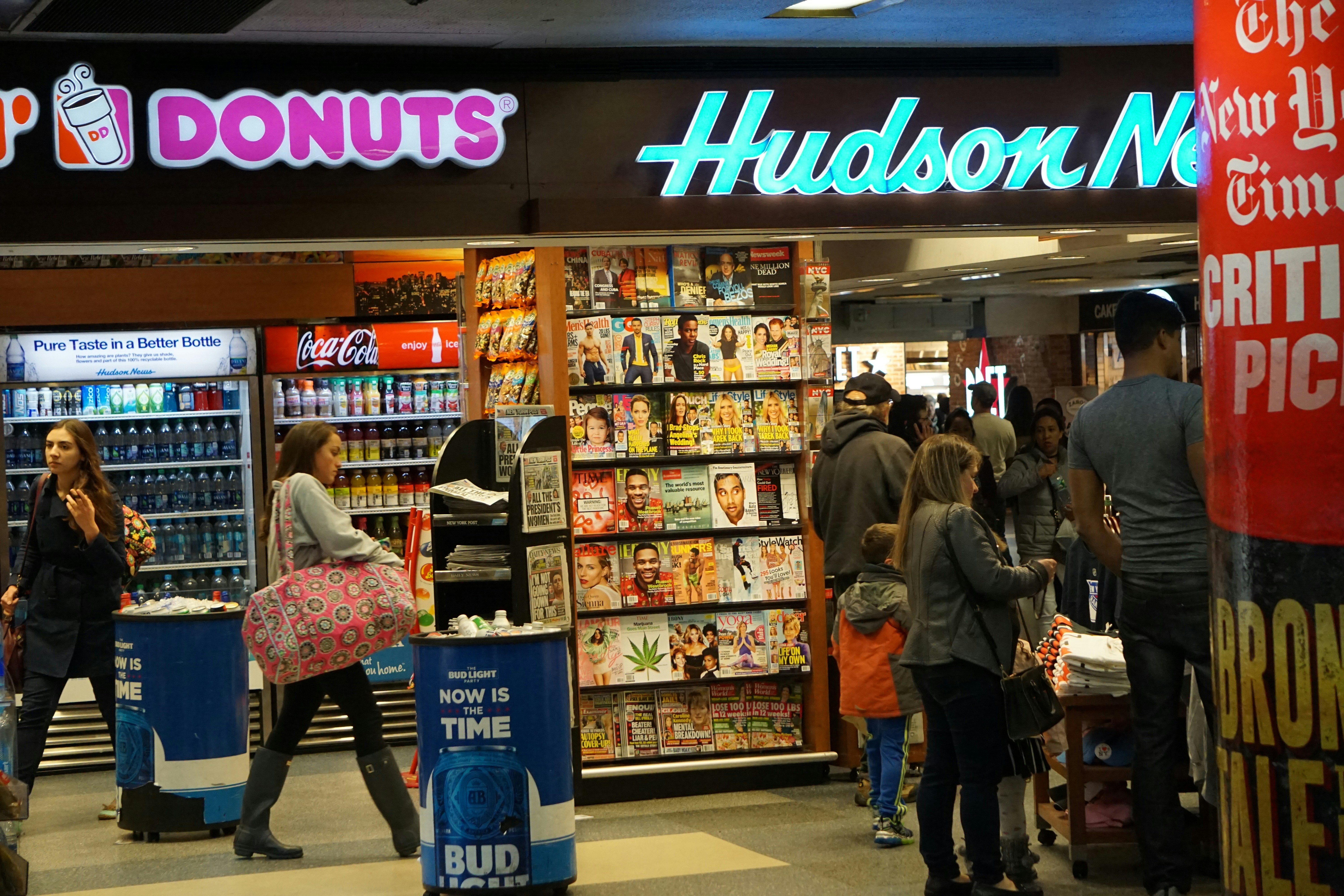 A busy newsstand with various magazines and newspapers on display. The scene includes people walking and browsing, with a Dunkin' Donuts and a Coca-Cola vending machine visible. Bright signage and numerous publications create a bustling and vibrant environment.